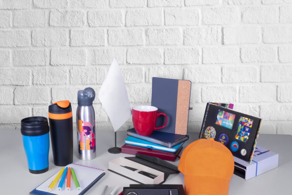 Composition of different promo products - Thermo mug, mug, gifts, pens in boxes, notebooks, tools, cap,flag table. Copy space. Grey wall background.